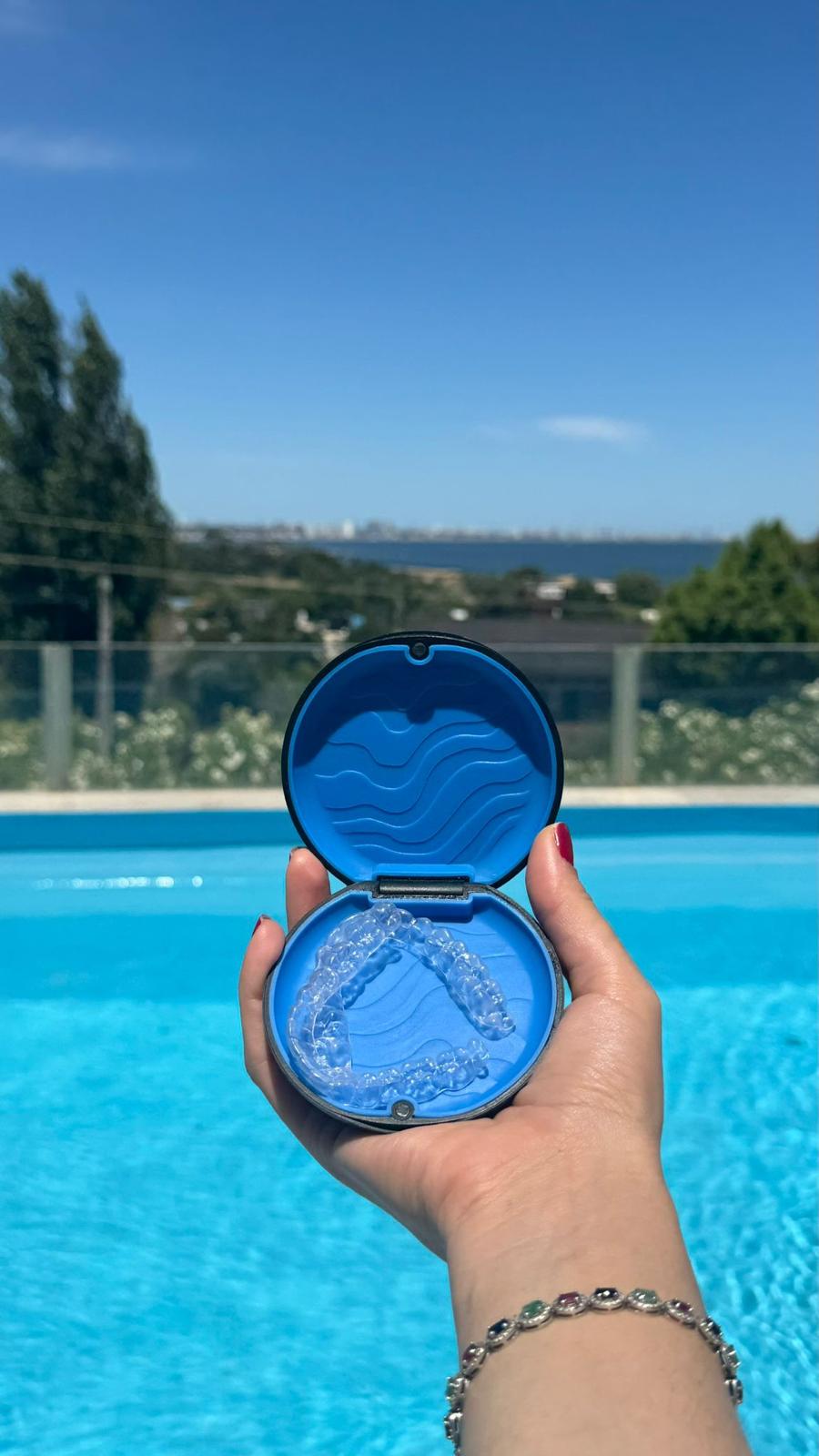 Mano sosteniendo el estuche azul de Sonrisa con los alineadores, frente a una piscina